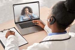 African doctor wear headset consult female black patient make online webcam video call on laptop screen. Telemedicine videoconference remote computer app virtual meeting. Over shoulder videocall view.