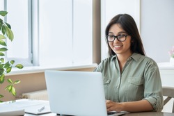 Happy indian young adult woman wearing glasses using pc laptop computer working studying at home office sitting at table. Happy female professional freelancer learning watching online webinar training