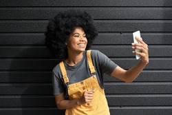 Happy African American hipster woman blogger with Afro hair laughing, holding phone, recording vlog, shooting social media stories or video calling in mobile app standing on black background.