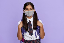 Indian preteen girl, latin kid schoolgirl student wears uniform and face mask for coronavirus protection safety holding backpack stands isolated on lilac violet background looking at camera, portrait.