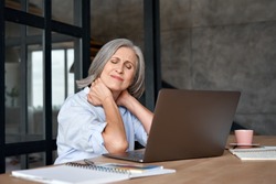 Tired stressed old mature business woman suffering from fibromyalgia neckpain working in office sitting at table. Overworked senior middle aged lady massaging neck feeling hurt pain from sedentary job