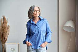 Smiling confident mature woman standing indoors looking at window. Stylish elegant middle aged senior 60s gray-haired lady thinking of good future vision, enjoying wellbeing, dreaming at home.