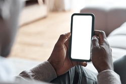 African american man holding smart phone with mockup white blank display, empty screen for app ads sitting on couch at home. Mobile applications technology concept, over shoulder close up view.