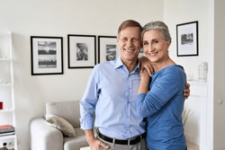 Happy senior mature 60s family couple hugging, looking at camera, standing in living room in modern apartment. Smiling satisfied middle aged husband and wife embracing posing for portrait at new home.