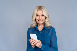 Happy middle aged business woman using apps looking at camera holding smartphone isolated on grey wall. Smiling 50s old lady advertising online mobile e bank services easy applications technology.