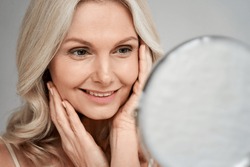 Happy 50s middle aged woman model touching face skin looking in mirror. Smiling mature older lady pampering, enjoying healthy skin care, aging beauty, skincare treatment cosmetic products concept.