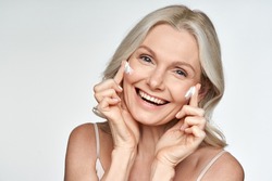 Happy 50s mid aged mature old blonde lady applying facial cream on face skin laughing enjoying anti age fresh soft skin care beauty spa treatment skincare creme isolated on white background. Portrait