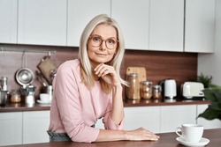 Happy mature middle aged 50s woman wearing glasses sitting at the kitchen table. Smiling older adult elegant blond lady looking at camera posing at home drinking coffee. Portrait.
