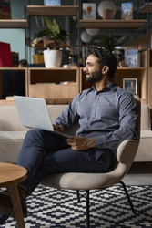 Young thoughtful bearded smart indian professional business man executive looking away sitting on chair in modern office lobby using laptop, thinking of new ideas, planning project, working online.