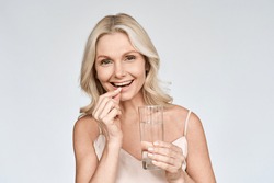 Happy middle aged 50s woman holding pill and glass of water taking dietary supplements. Portrait of smiling adult attractive woman taking care of health in menopause, isolated on white.