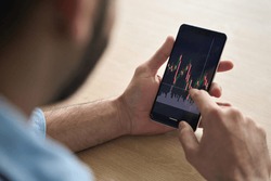 Over shoulder close up view of male analyst broker's hands holding touch screen device smartphone using tech ecommerce application of finance stock markets with graphs and numbers.