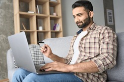 Young attractive indian Hispanic male consumer doing online virtual shopping using laptop and holding credit debit card sitting on sofa at home. Ecommerce and secure financial payments concept.