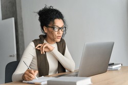 Young adult African American mixed race student wearing glasses watching online webinar on pc writing notes on desk at home modern home office. Remote distant e learning work concept.