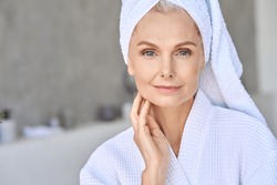 Happy smiling attractive senior older woman wearing bathrobe and white towel touching face looking at camera at bath room. Advertising of skin care spa procedures concept. Closeup portrait.