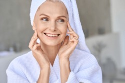 Portrait of happy attractive middle aged woman wearing bathrobe and white turban with bright complexion touching face looking away in bathroom. Advertising of skin care spa wellness concept.