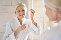 Senior mature older caucasian woman touching clean face eye contour with antiaging pipette serum essence oil looking at mirror wearing bathrobe. Anti wrinkle prevention skin care products concept.