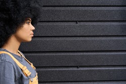 Young beautiful black hipster girl of 20s standing side view profile on black wall background. Headshot of stylish serious funky girl with afro hair looking at copy space.