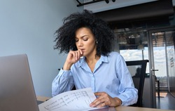 Concerned African American businesswoman ceo analyst sitting at desk reading papers financial reports with laptop in contemporary corporation office. Financial data analyst working with big data.