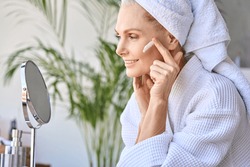 Gorgeous senior older adult 50 years old blonde woman wearing bathrobe and turban towel in bathroom applying moisturizing tightening face skin treatment, looking at mirror. Morning beauty routine.
