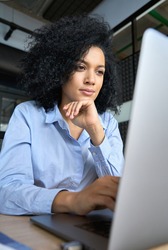 Young serious concerned African American businesswoman sitting at desk looking laptop computer in contemporary corporation office. Business technologies concept. Vertical portrait.
