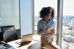 Young focused African American businesswoman sitting on desk holding using tablet computer device in modern office in high floor building with panoramic urban capital city view.