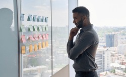 Serious thoughtful young indian businessman ceo near whiteboard with adhesive sticky notes thinking about ideas of planning strategy project standing at panoramic window of modern business office.