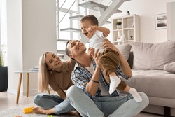 Happy young family couple having fun playing with cute small toddler kid son in modern living room together. Smiling parents mum and dad enjoying spending time with cute funny child boy at home.