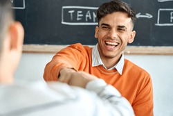 Young happy laughing Indian latin Hispanic creative startup coworker student giving fist bump to classmate partner committing successful project result in office classroom at desk near blackboard.