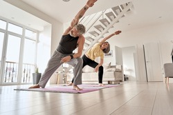 Fit middle aged 50s family couple doing fitness yoga morning exercise at home. Sporty healthy old mature man and woman training together standing in living room. Active seniors sport stretching.