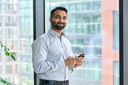 Smiling indian ceo businessman holding using cell phone mobile apps standing in office near panoramic window looking at camera. Digital tech apps and solutions for business corporate development.