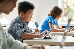 Focused Asian school boy using digital tablet at class in classroom. Attentive junior school student learning online virtual education digital program app tech during stem computer science lesson.