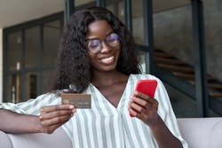 Happy young black African woman customer shopper holding credit card using cell phone mobile app buying fashion clothes paying online making purchase in ecommerce digital store on smartphone at home.