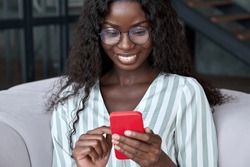 Young happy black African woman wearing glasses using mobile digital apps on cell phone tech device for online shopping or dating sitting on sofa at home, texting on smartphone, writing social post.