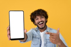 Happy excited young indian man showing smartphone pointing at big mockup white blank phone template screen isolated on yellow background presenting cellphone mobile offer application ads concept.