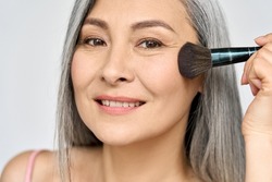 Senior older middle aged Asian woman with grey hair holding make up brush on radiant face with perfect skin. Advertising of radiant foundation skincare and makeup for natural glow and healthy skin.