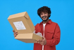 Happy indian young man holding open parcel box isolated on blue background. Smiling guy customer receiving product retail order purchase in postal shipping delivery unpacking package.