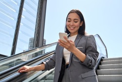 Smiling young adult Asian business woman wearing suit standing on urban escalator using applications on cell phone gadget, reading news on smartphone, browsing fast mobile internet outdoors.