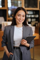 Young smiling successful professional leader Asian business woman, female executive manager, saleswoman wearing suit holding digital tablet standing in office looking at camera, vertical portrait.
