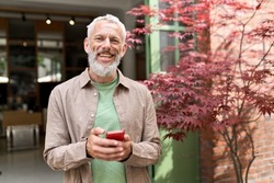 Smiling gray-haired older middle aged bearded man using mobile phone outdoors. Happy old senior adult male user holding cellphone texting on smartphone looking at camera standing outside.