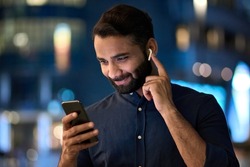 Smiling bearded indian man wearing earbud holding phone having video call at night. Eastern businessman in earphone using smartphone listening music in app tech on cellphone watching videos online.