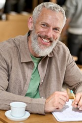 Happy older adult business man professor or student writing in notebook, smiling middle aged gray-haired bearded author or writer taking notes looking at camera sitting at table. Vertical portrait.