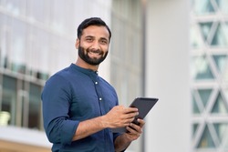 Smiling young adult indian business man professional executive holding digital tablet online fintech outside, happy eastern businessman ceo using pad standing outdoors in big urban city street.