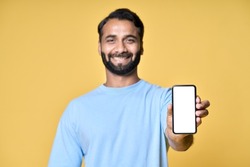 Happy indian man showing cellphone pointing at big mockup white blank phone template screen isolated on yellow background presenting smartphone mobile shop offer ads. Betting marketing concept.