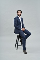 Happy successful rich young indian business man ceo leader, wealthy arab professional manager, confident male businessman executive wearing suit sitting on chair isolated on beige, vertical portrait.