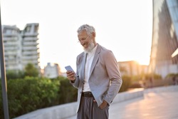 Happy older mature adult professional business man, smiling senior old businessman wearing suit holding smartphone using mobile cell phone standing outdoor in big city downtown among office buildings.