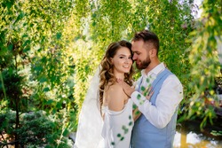 groom in blue suit and bride with long curls, in a white dress with open shoulders and a bouquet of lilies stand in willow. wedding photo shoot in the park.
