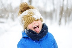 Funny little boy in blue winter clothes walks during a snowfall. Outdoors winter activities for kids. Cute child wearing a warm hat low over his eyes catching snowflakes with his tongue