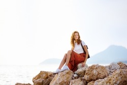 Charming young woman sitting on the Mediterranean coast. Attractive red-haired girl sits on rocks by the sea
