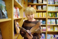 A preteen boy leafing through a book while standing at the bookshelfs in a school library or bookstore. Smart kid reading comics or adventure book