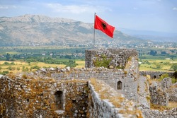 Red Albanian flag with double-headed black eagle waving over wall of Fortress Rozafa near Shkodra city. Fluttering banner with symbol of Albania on background of sky. Travel and tourism in Balkan